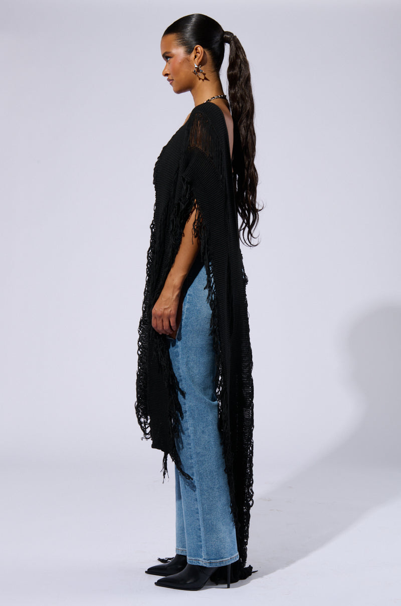 A person with long, dark hair styled in a high ponytail stands in profile against a plain background. They are wearing the "GOOD VIBES DISTRESSED PONCHO IN BLACK" with wide sleeves, blue jeans, and black pointed-toe boots—a true Boho Chic Ensemble.
