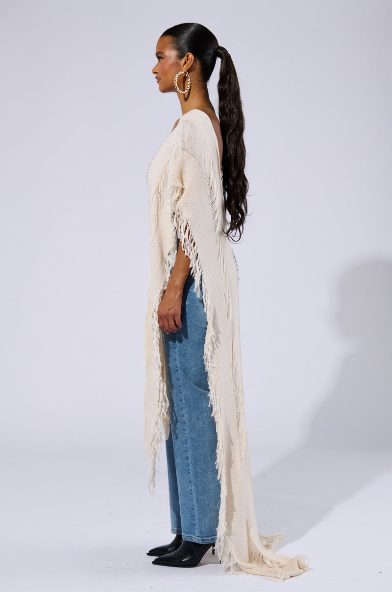 Profile view of a woman with long, dark hair tied in a high ponytail, wearing large hoop earrings, the GOOD VIBES DISTRESSED PONCHO IN CREAM, blue wide-leg jeans, and black heels. She is facing left against a plain background.