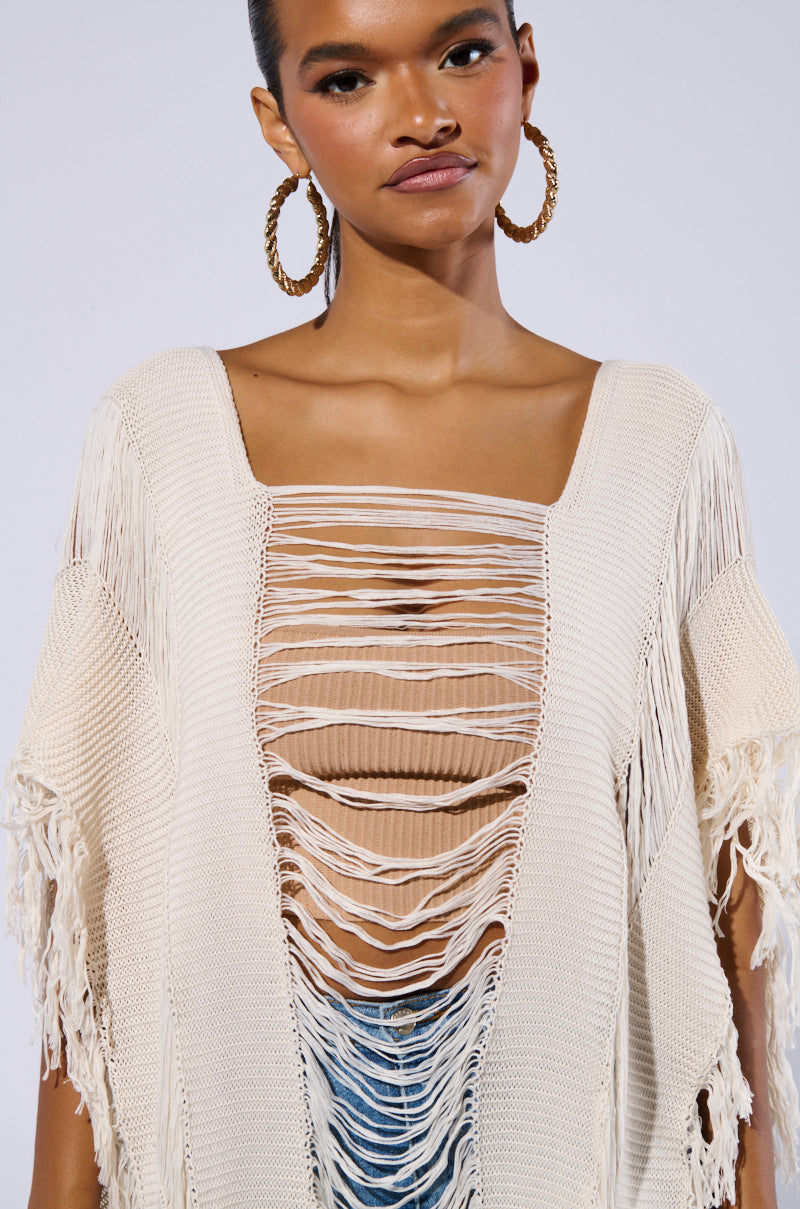 A woman is wearing large hoop earrings and the GOOD VIBES DISTRESSED PONCHO IN CREAM, which features a light beige, crochet knit design with a rectangular cutout pattern on the front. The poncho reveals a ribbed beige tank top underneath. Sporting short-cropped hair, she poses against a plain, light background, evoking a chic festival ensemble vibe.