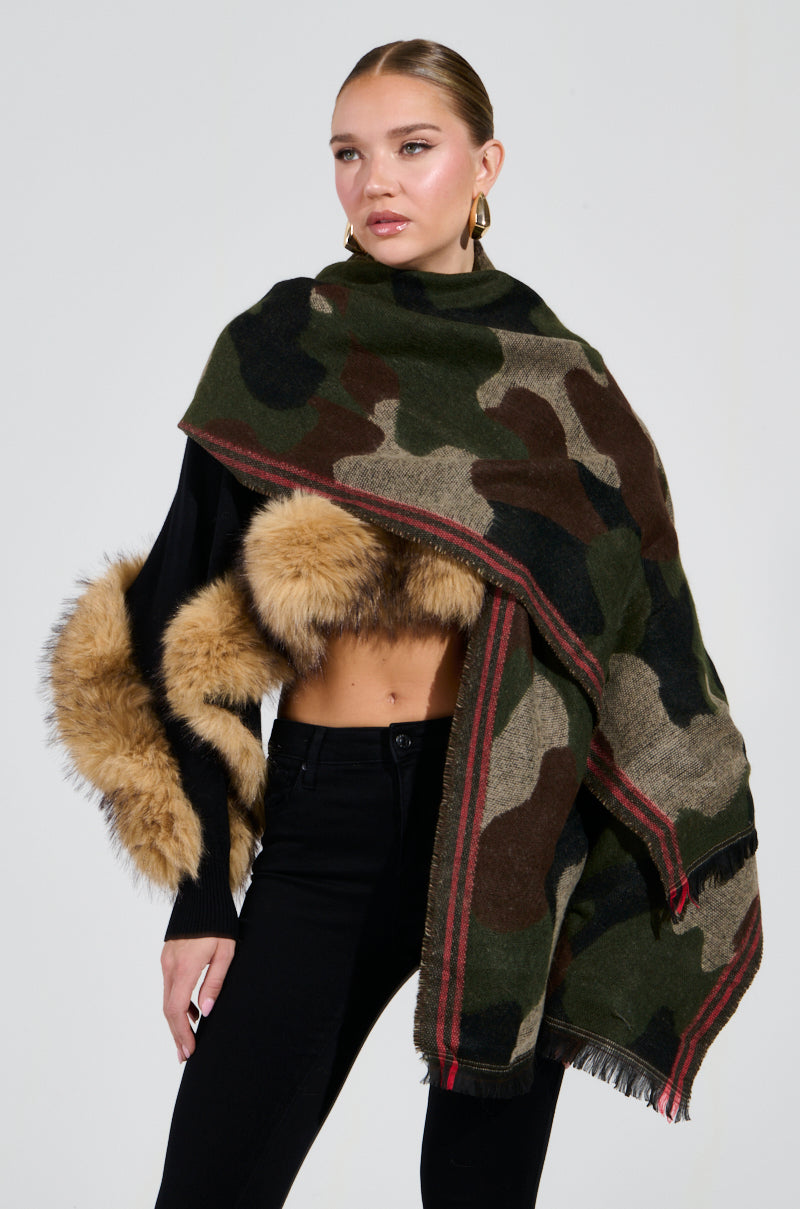 Draped elegantly over her shoulders, a woman in a sleek black outfit showcases the oversized silhouette of the CAMO NIGHTS SCARF. The fur-trimmed scarf enhances her chic appearance as she poses against a plain background, with her hair pulled back.