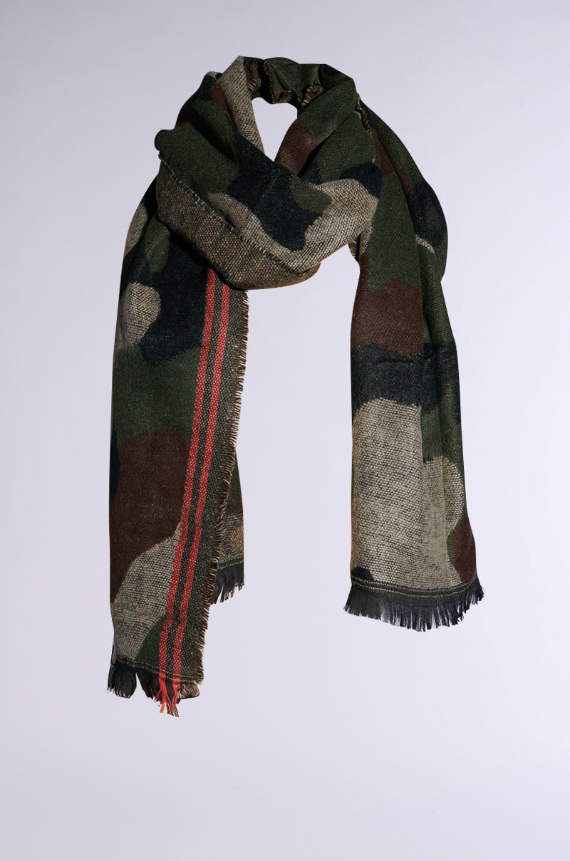 The CAMO NIGHTS SCARF boasts a camouflage design in hues of green, brown, and black, complemented by bold red stripes on one side and finished with black fringes at the ends. Its oversized knit silhouette is perfect for styling in a loose knot against a simple light backdrop.
