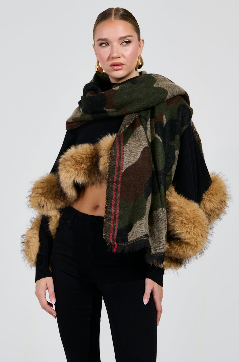 A woman showcases a stylish ensemble highlighted by the CAMO NIGHTS SCARF, characterized by its oversized silhouette and draped over a black top with fur cuffs. She completes the look with black pants, neatly styled hair, and gold hoop earrings for an effortlessly chic appearance.