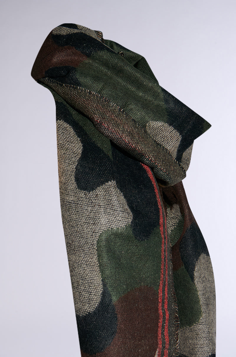 A close-up of the CAMO NIGHTS SCARF, showcasing its green and brown camouflage-patterned fabric draped over a surface. This cozy knit fabric features a distinct red stripe running along one edge, with a textured appearance and visible folds.