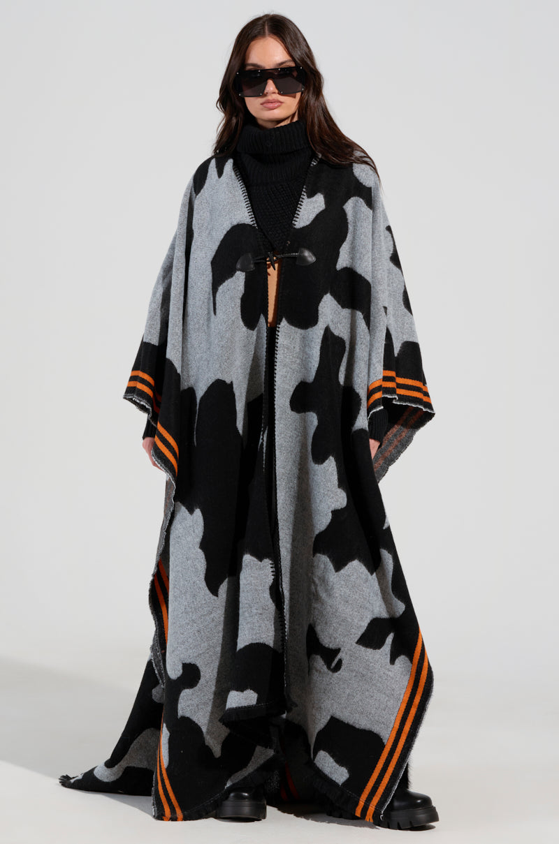 A person wearing oversized sunglasses and the striking MOD LIFE CAMO PONCHO, featuring a black and gray abstract pattern with orange stripe accents at the edges. The ultra-soft knit poncho drapes elegantly to the floor as the person stands confidently against a plain background.