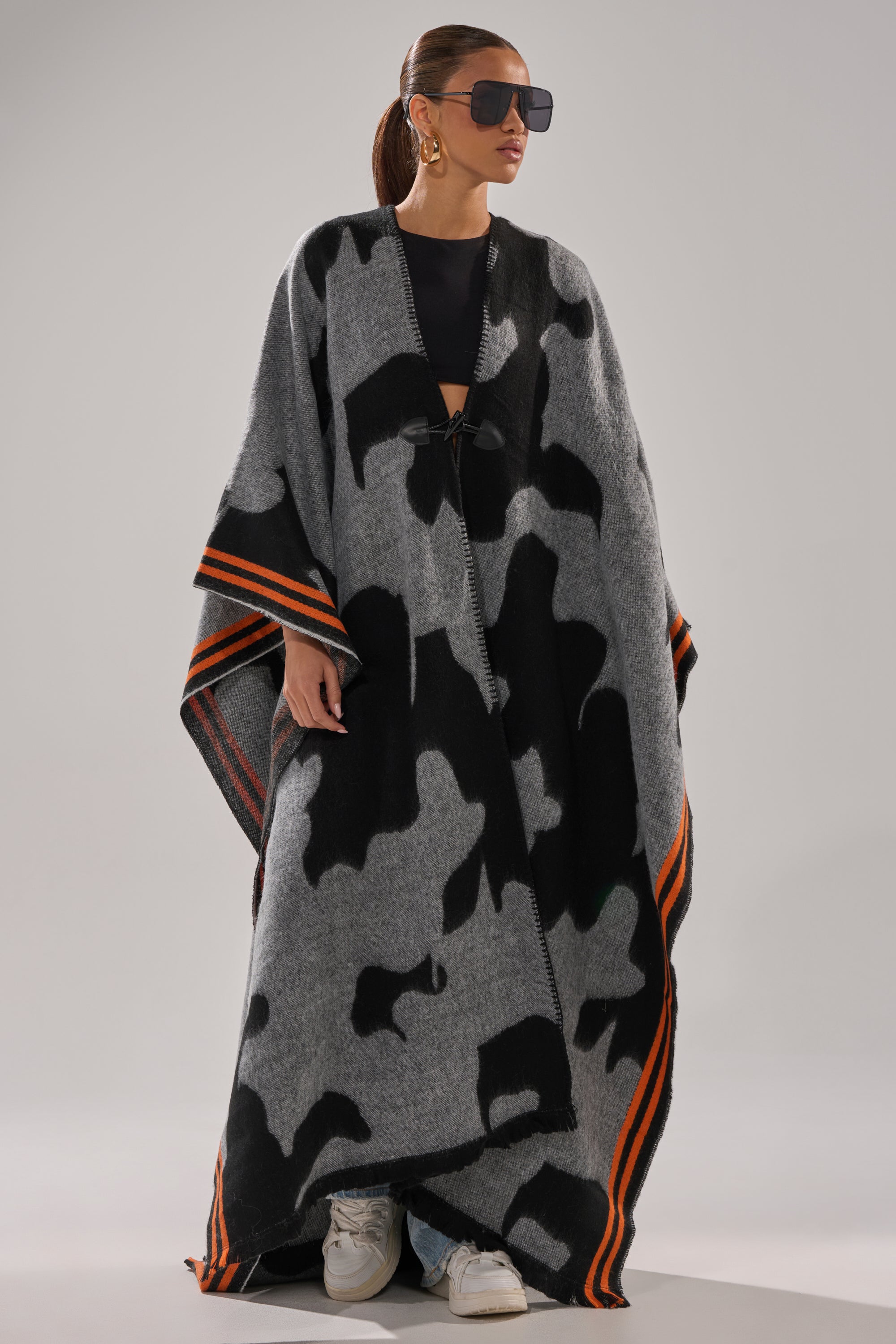A woman models the MOD LIFE CAMO PONCHO—maxi length with gray and black patterns and orange striped edges—over a black outfit, paired with oversized sunglasses, hoop earrings, and white sneakers.