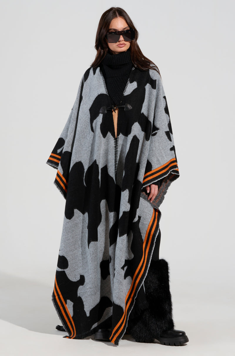 A person models the MOD LIFE CAMO PONCHO, featuring an ultra-soft knit with gray and black camouflage patterns and orange trim, paired with sunglasses and black fur boots, against a simple backdrop.