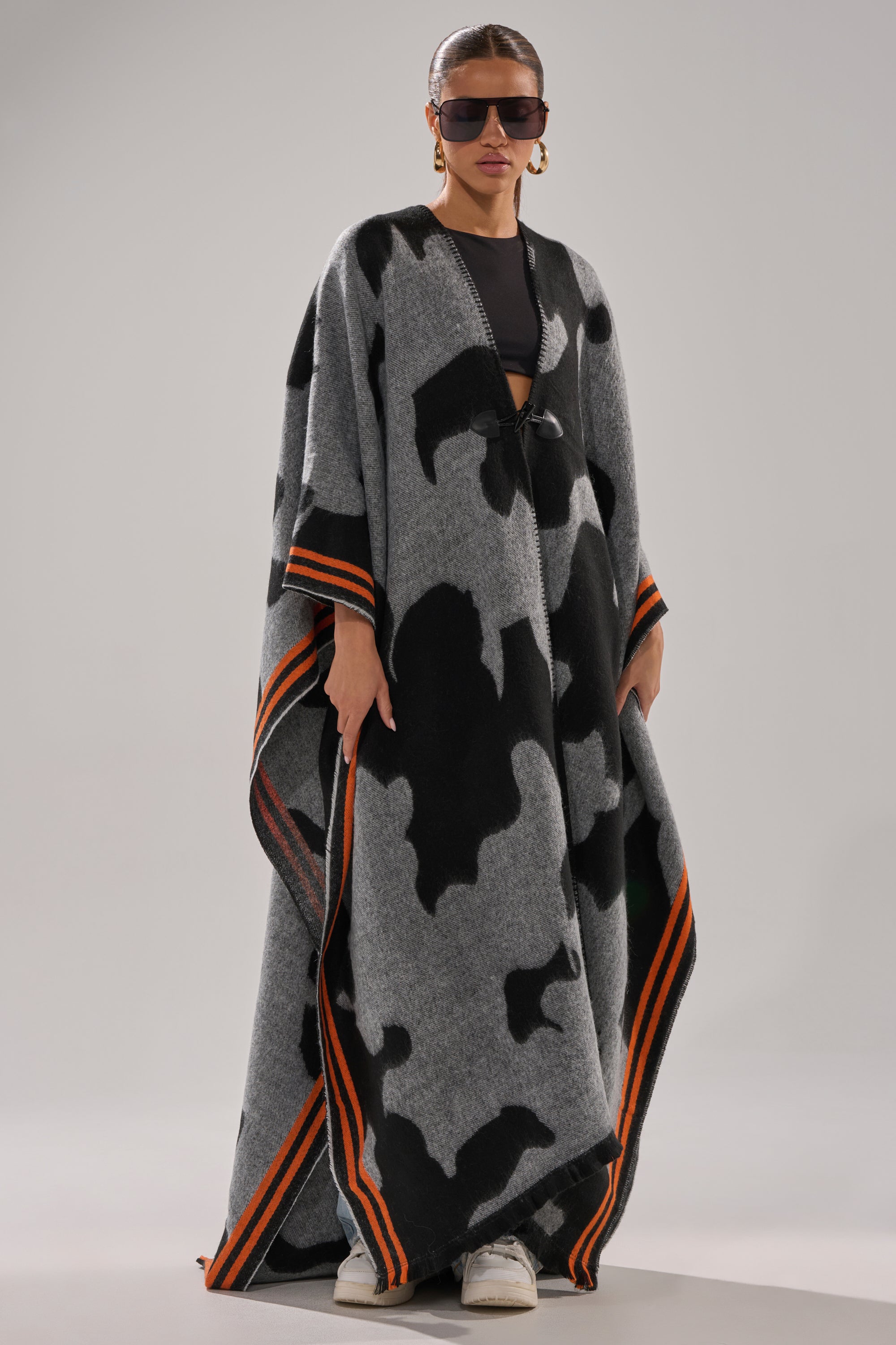 A woman models the MOD LIFE CAMO PONCHO, a long gray and black abstract-patterned piece with orange-striped edges, paired with oversized sunglasses and gold earrings, over a black outfit and white shoes against a plain background.