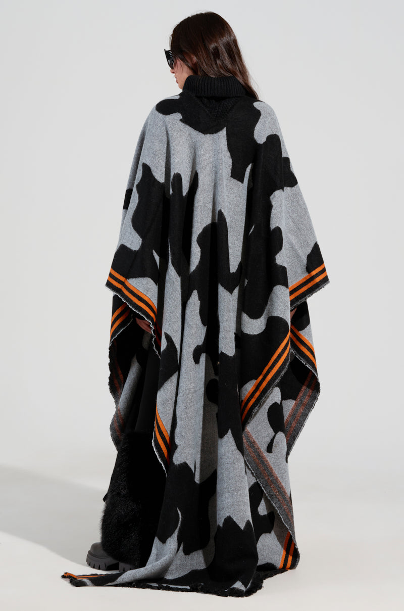 A person is dressed in the MOD LIFE CAMO PONCHO, which features a black, white, and gray camo pattern with orange striped edges. They have long, dark hair and are wearing sunglasses while facing away from the camera.