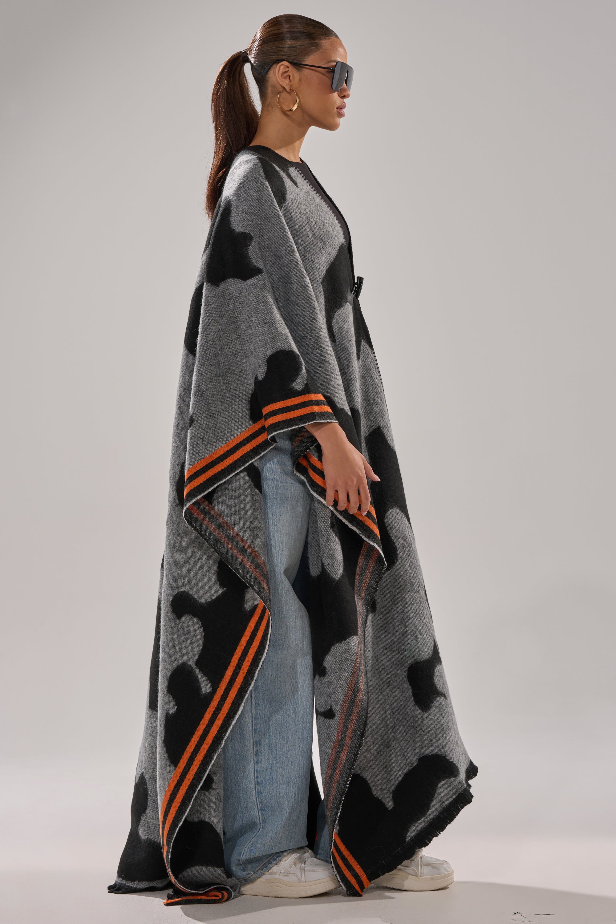 A woman in sunglasses stands sideways against a light background wearing the MOD LIFE CAMO PONCHO—a gray and black knit poncho with orange stripes—styled over blue jeans and white sneakers.