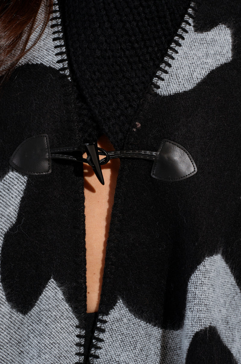 Close-up of the MOD LIFE CAMO PONCHO in black and gray, featuring a toggle clasp. The ultra-soft knit fabric showcases a bold, abstract pattern, and the clasp is crafted from leather.