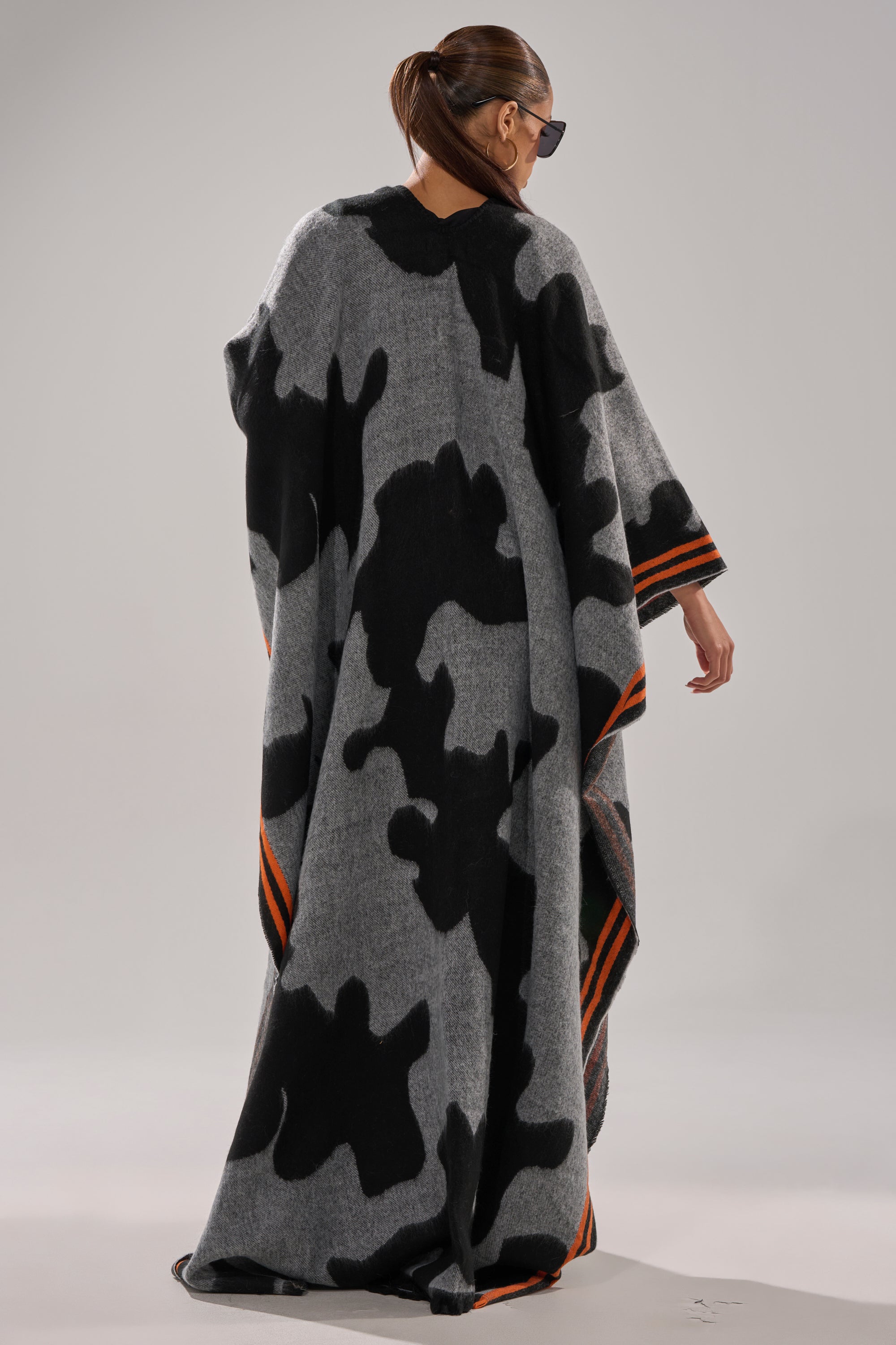 A person with long brown hair in a ponytail, wearing sunglasses and the MOD LIFE CAMO PONCHO featuring orange stripes near the hem, stands facing away in a minimalist studio setting.