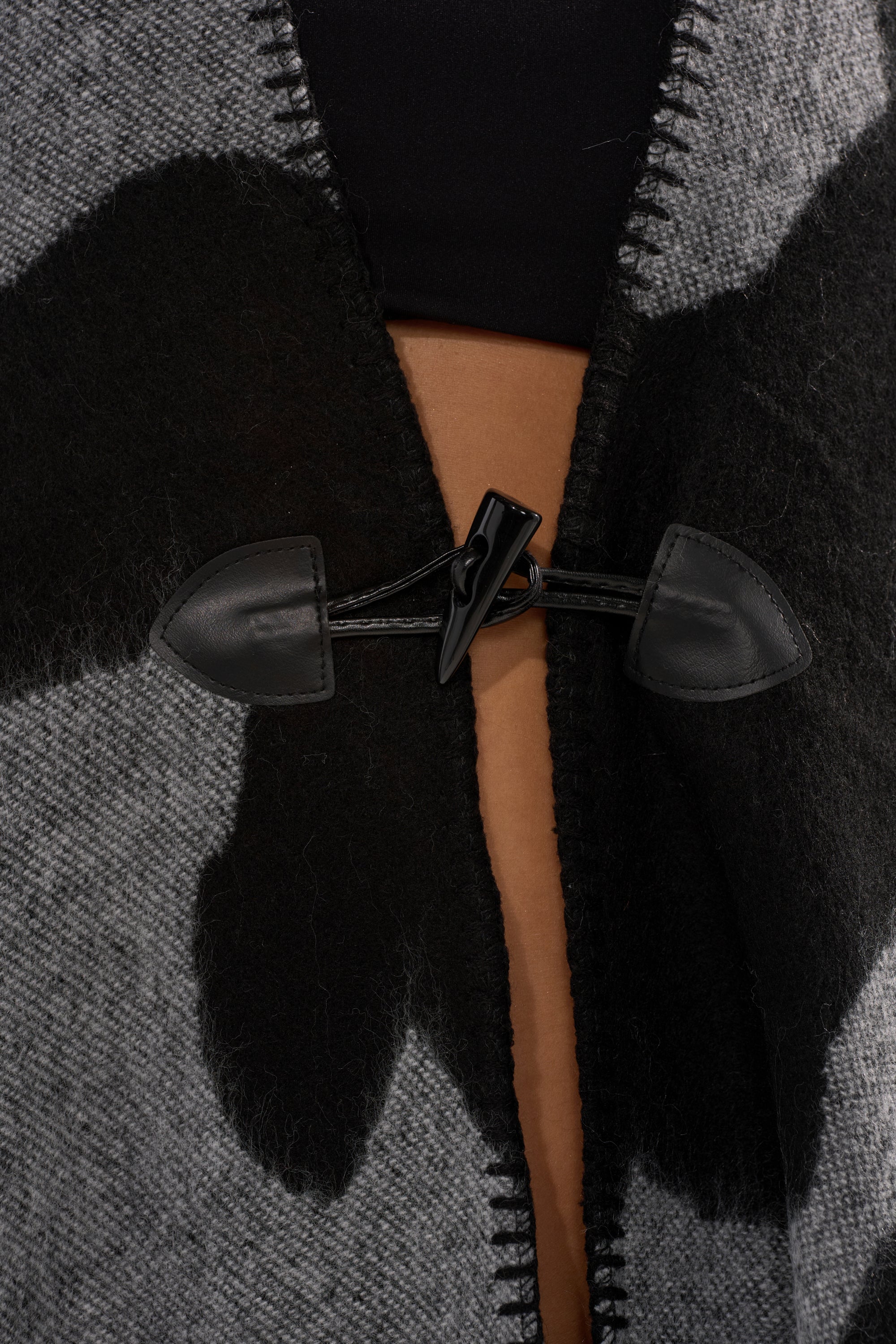 Close-up of the toggle clasp fastening on the MOD LIFE CAMO PONCHO, featuring thick black and gray wool. Partially open to reveal bare skin and a black top, this poncho highlights visible stitching and rich fabric texture—perfect for cozy wardrobes.