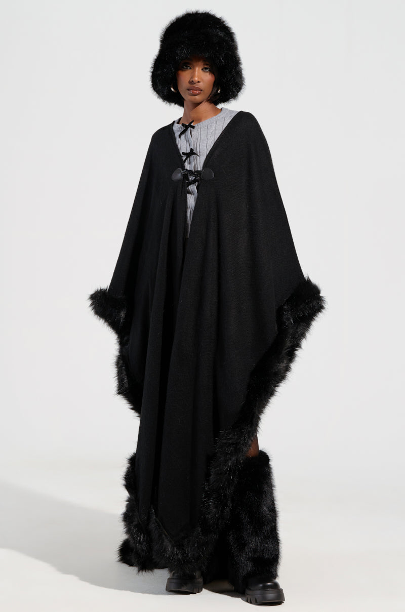 A person dressed in a winter-ready outfit with an oversized silhouette, showcasing the LOVE MY LIFE FAUX FUR TRIM PONCHO in black, paired with a matching fur hat and boots. Underneath, a light-colored ensemble provides a beautiful contrast to the plain and bright background, accentuating the striking dark attire.
