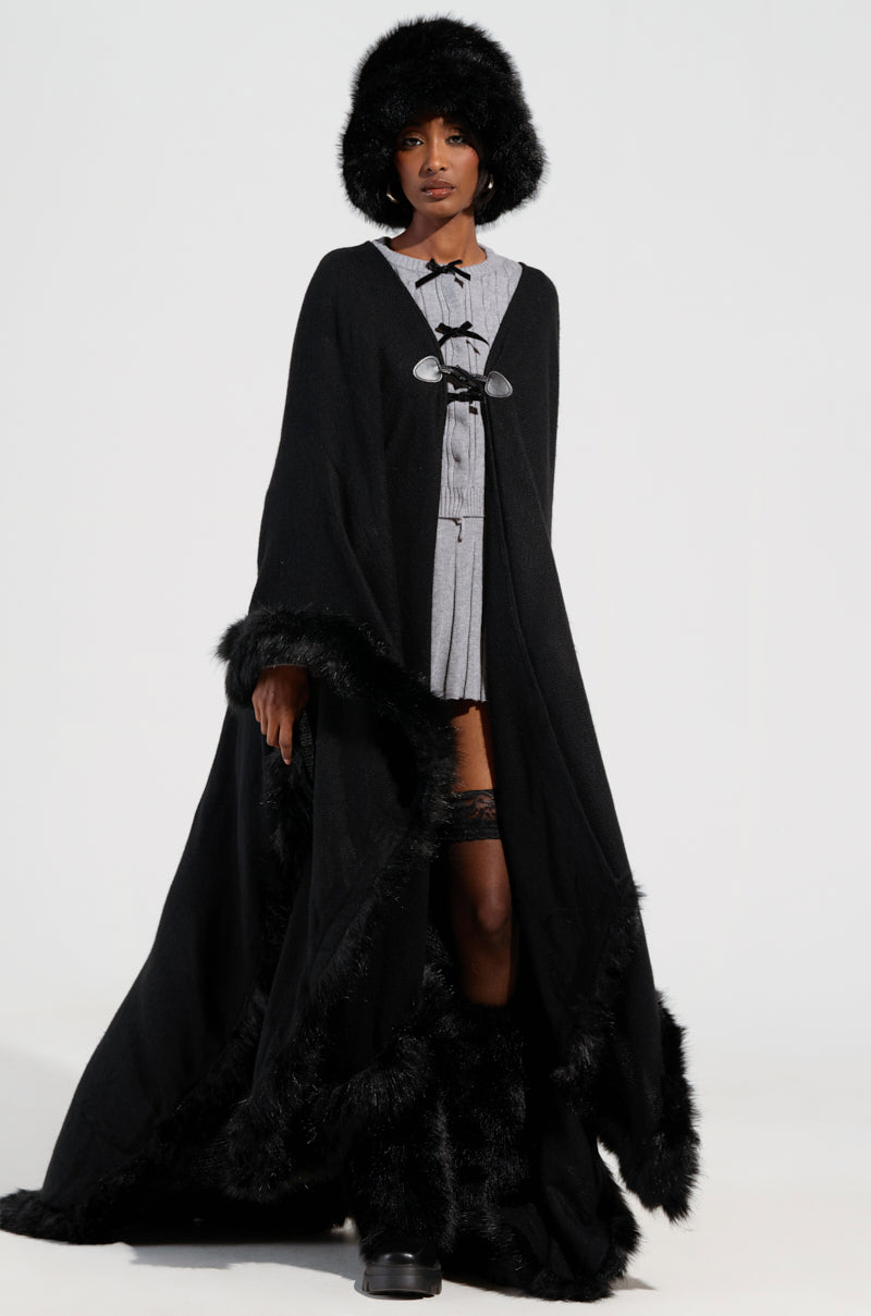 A person is winter ready in an oversized silhouette, wearing the striking LOVE MY LIFE FAUX FUR TRIM PONCHO and matching hat. Beneath, a gray top complements high boots. The poncho elegantly reveals part of their leg against the plain light-colored background.