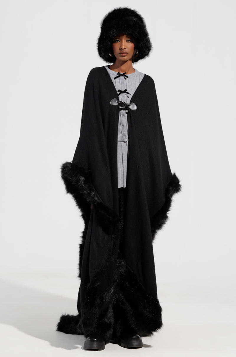 A person stands dressed in a gray shirt under the LOVE MY LIFE FAUX FUR TRIM PONCHO, featuring an oversized black cloak design with faux fur trimming. Their outfit, paired with a fluffy black hat and large boots, is perfectly suited for winter against the plain white background.