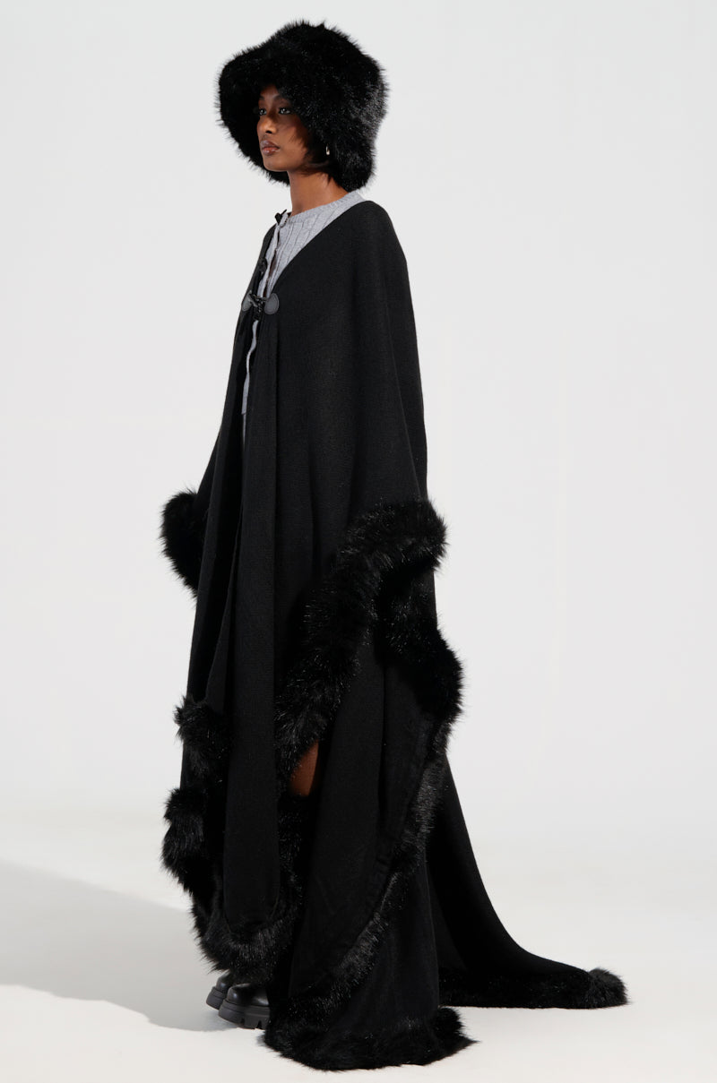 The LOVE MY LIFE FAUX FUR TRIM PONCHO is showcased in a winter-ready fashion, featuring a long black cape with faux fur trim and a matching hat. The ensemble, complete with a high collar and button detail accented in gray, presents an oversized silhouette against a plain white background.