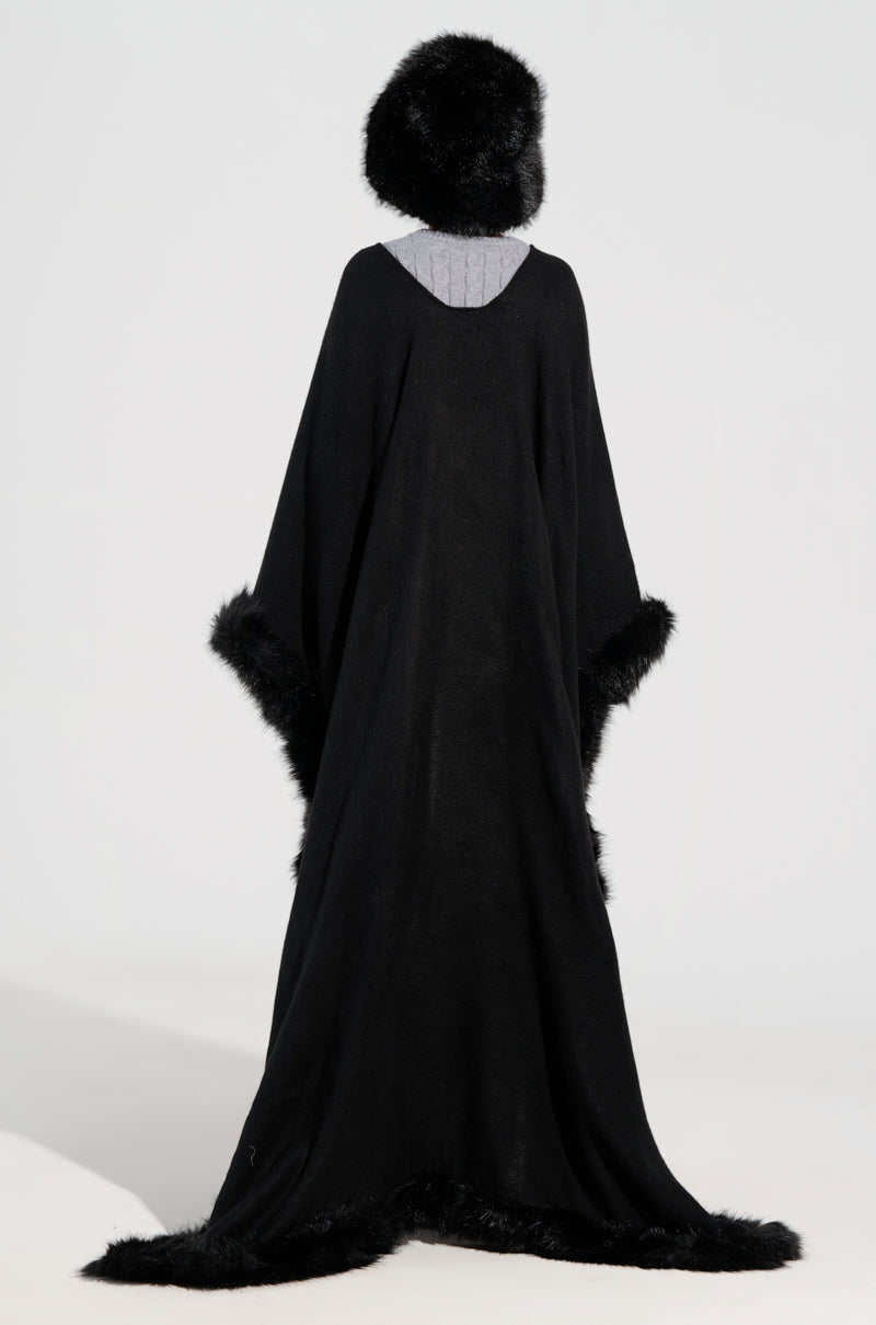 Dressed in an oversized silhouette, the individual dons the LOVE MY LIFE FAUX FUR TRIM PONCHO—a voluminous black cloak with a luxurious faux fur trim—and pairs it with a matching fur hat. They stand against a plain grey backdrop, turned away from the camera, showcasing winter-ready elegance.