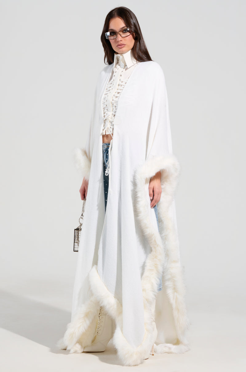 Someone draped in the LUXE LIFE FAUX FUR TRIM PONCHO, over a chic blouse and jeans. They carry a compact handbag and sport sunglasses, showcasing an oversized silhouette against a minimalist background.
