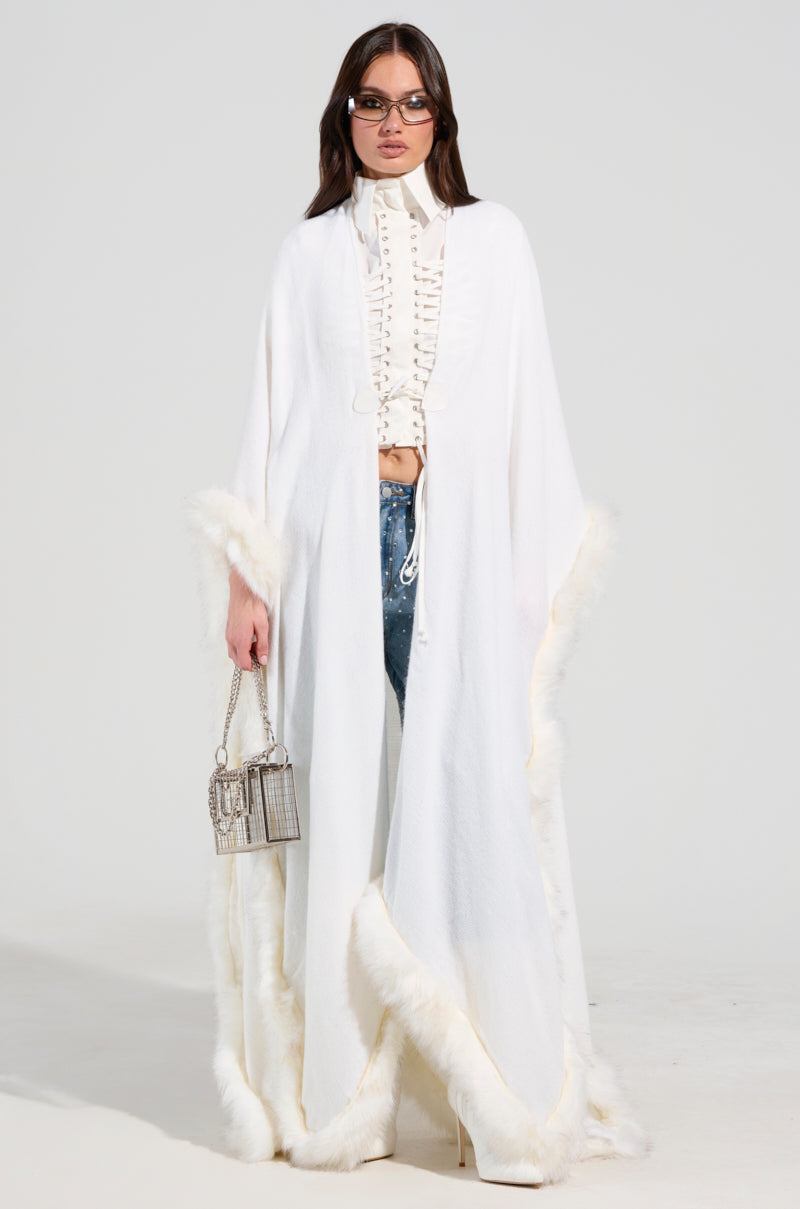 A person dressed in a stylish ensemble showcases the oversized silhouette of the LUXE LIFE FAUX FUR TRIM PONCHO, complemented by a laced blouse and sunglasses. They carry a small metallic handbag, creating a chic, winter-ready look against the plain white background.