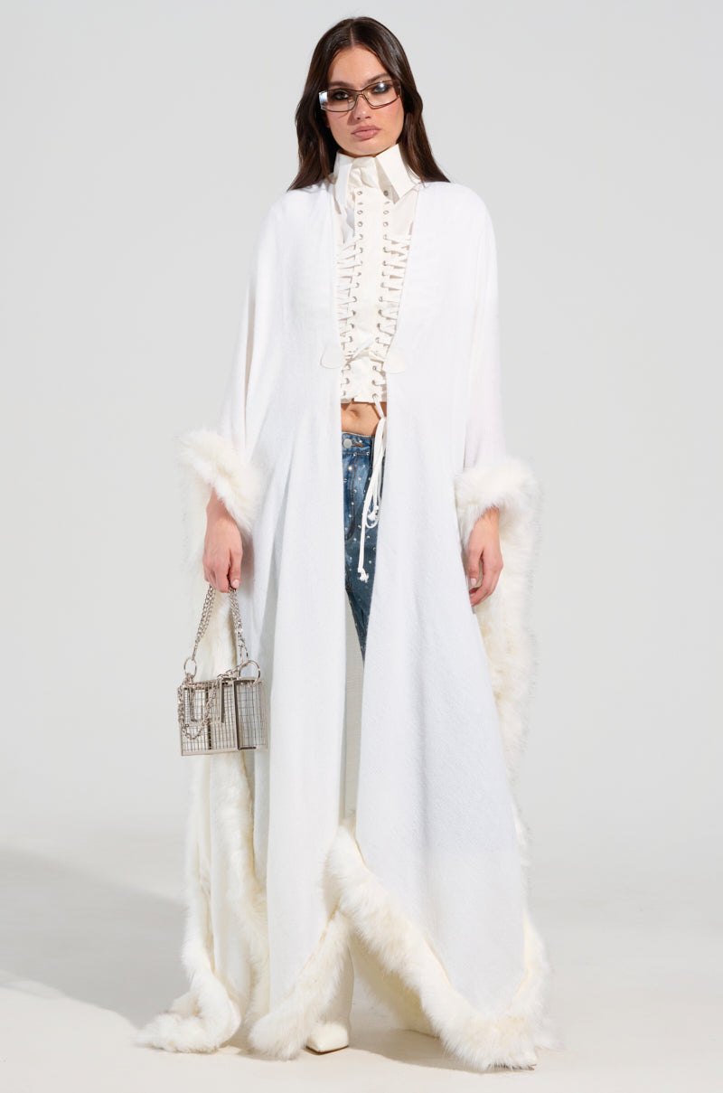 A woman effortlessly embodies Luxe Life, draped in a LUXE LIFE FAUX FUR TRIM PONCHO over a ruffled shirt and denim jeans. She accessorizes with sunglasses and a small metallic handbag, exuding a winter-ready finish against the plain background.