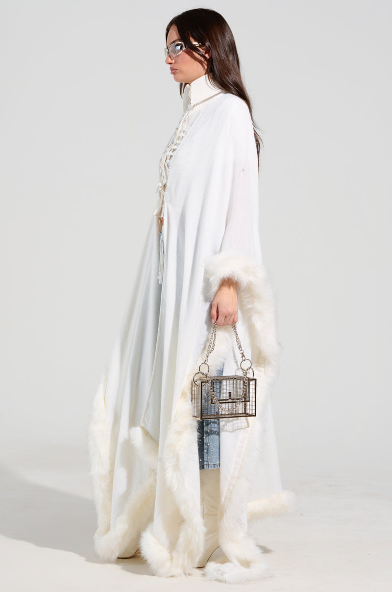 Dressed in the LUXE LIFE FAUX FUR TRIM PONCHO, a person stands in profile wearing glasses against a plain background. Their luxurious white cloak is complemented by a decorative, cage-like handbag and platform shoes, creating an elegant winter-ready look.