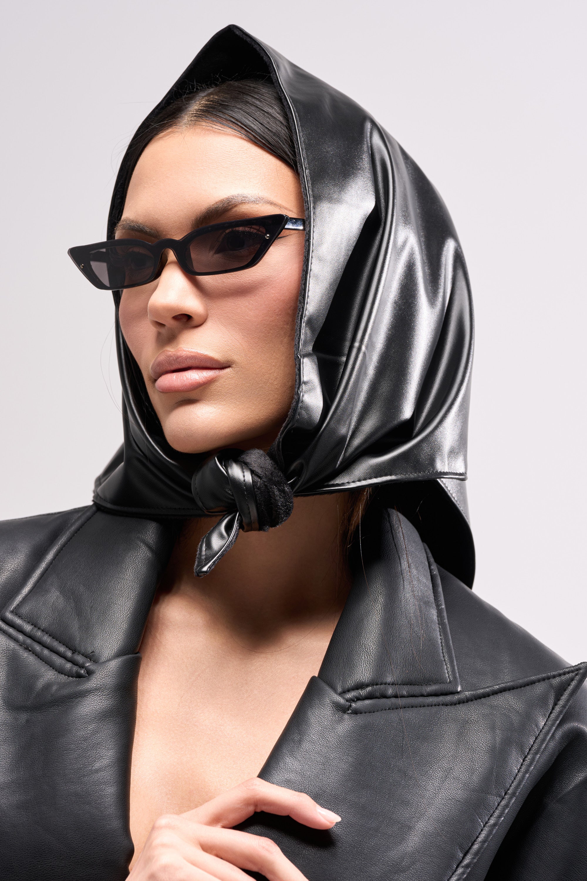 A woman gazes confidently to the side against a plain light background, wearing the ITS A WRAP PU HOOD, cat-eye sunglasses, and a black AKIRA Label faux leather jacket.