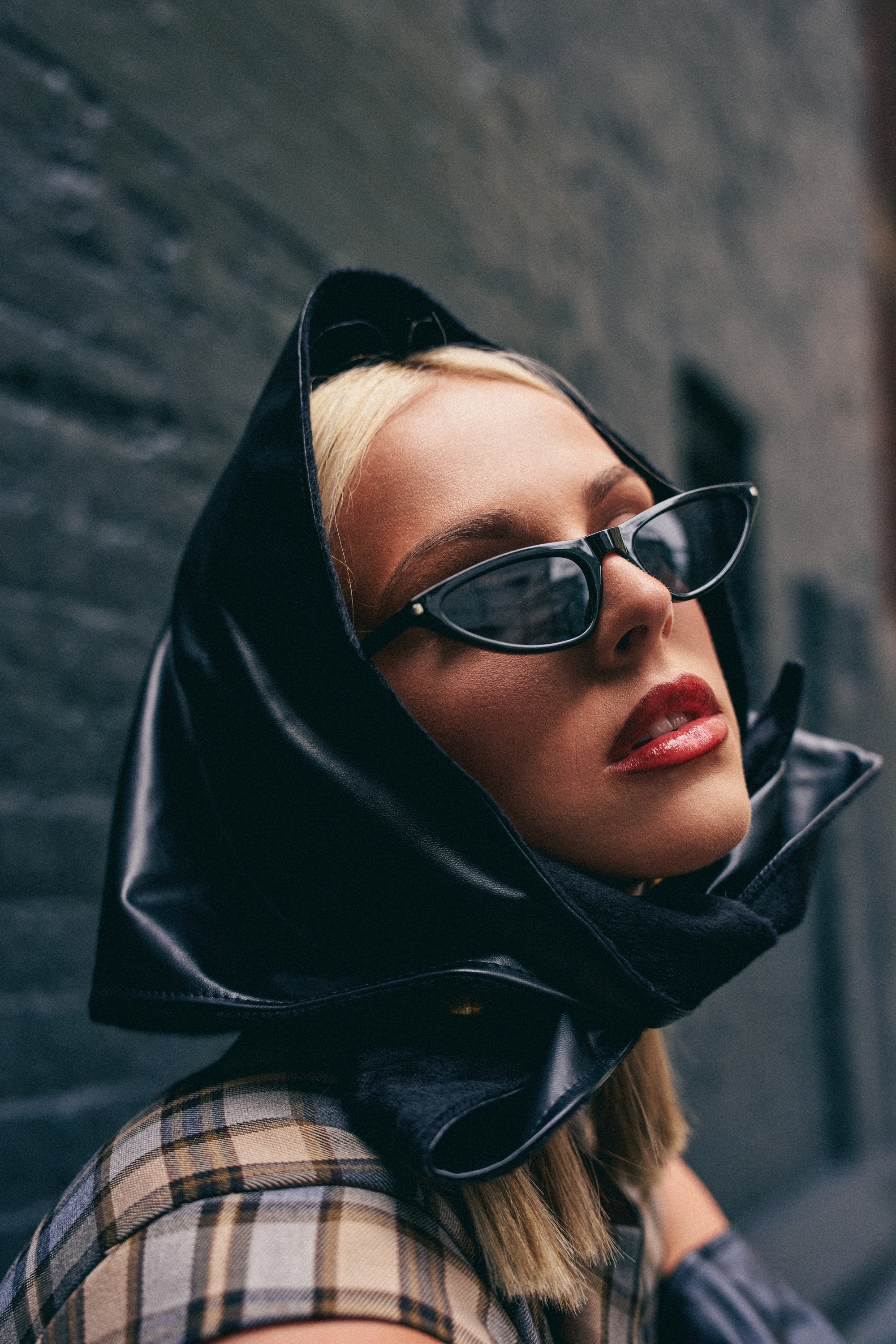 Wearing the ITS A WRAP PU HOOD draped stylishly over her head, along with cat-eye sunglasses and a plaid coat, a woman poses confidently against a textured gray wall, perfectly capturing modern streetwear style.
