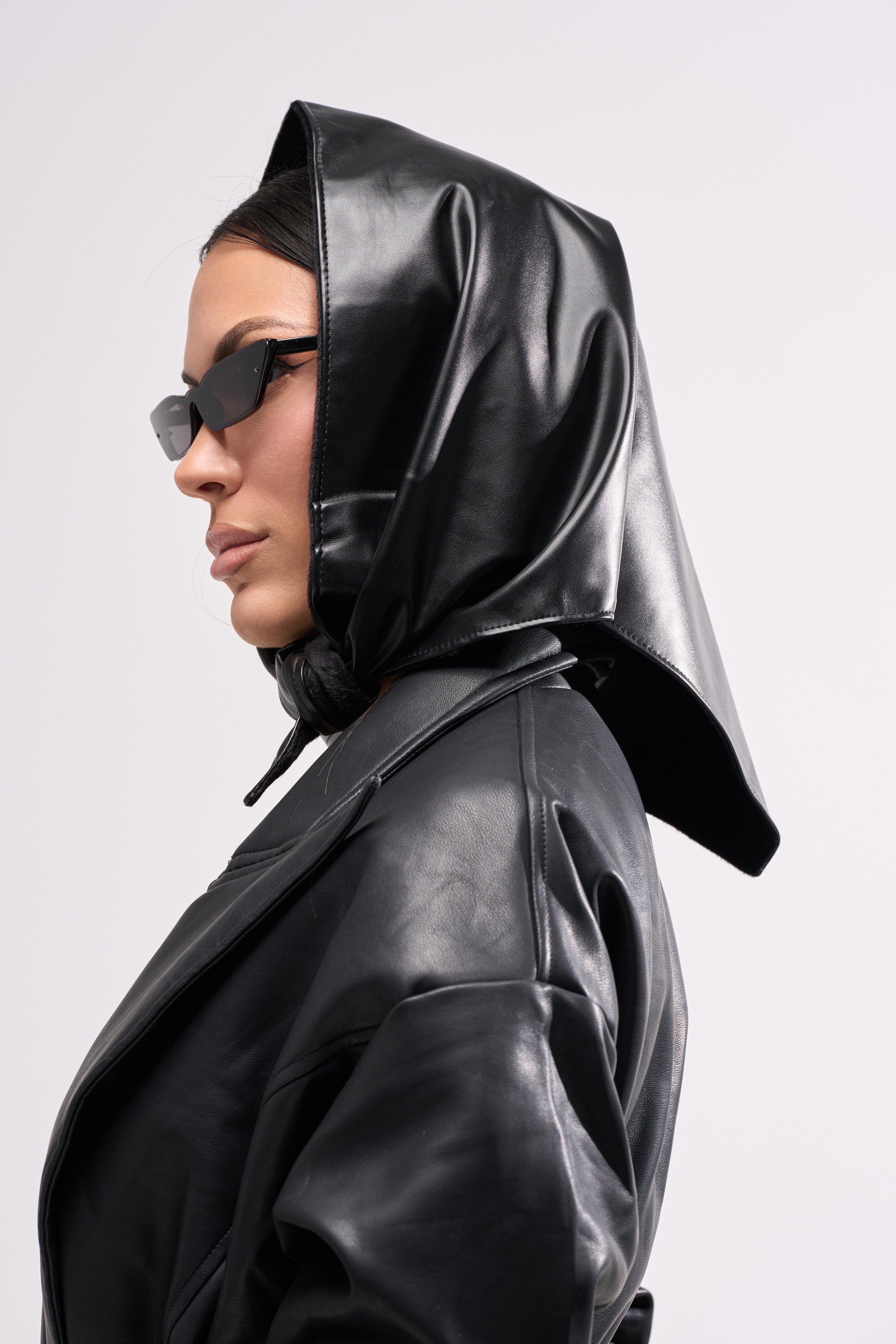 A woman wearing the ITS A WRAP PU HOOD, black sunglasses, and a black leather jacket from AKIRA Label is shown in profile against a plain light background. 1