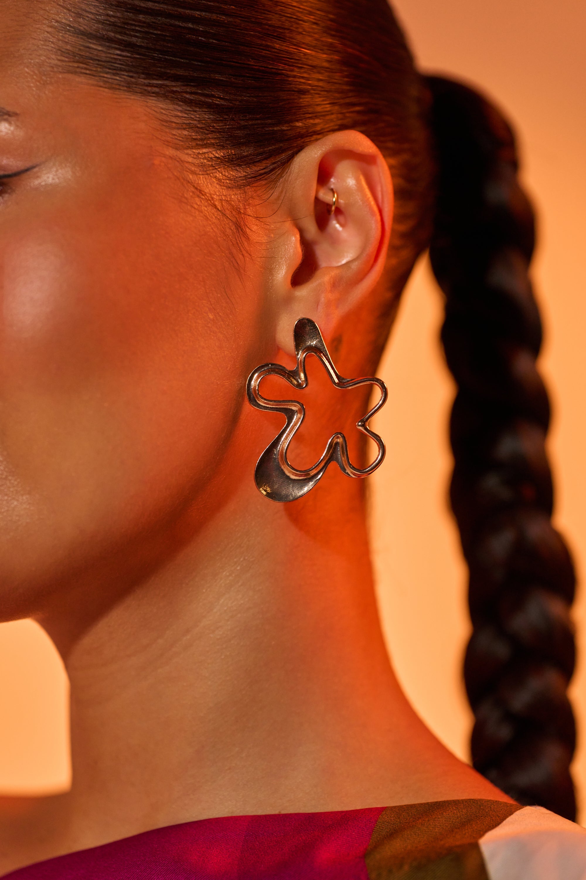 A close-up of a woman's ear wearing the VIC EARRING—a large, gold-tone, floral-inspired statement piece. Her dark hair is styled in a sleek braid against a softly lit background with warm orange tones. 0