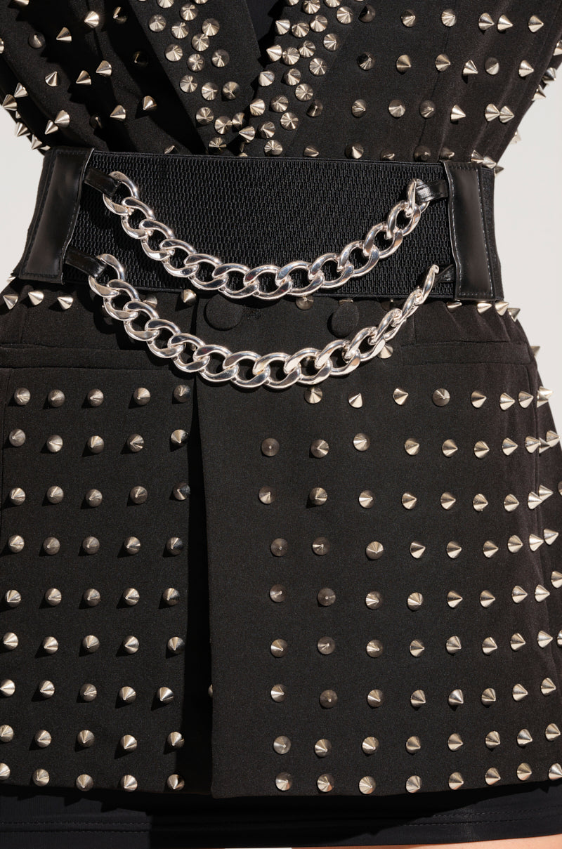 A close-up of a person wearing a black studded jacket with silver spikes, cinched at the waist with THE REGAL CHAIN BELT—a wide black elastic belt adorned with two hanging faux silver metallic Cuban chain links.