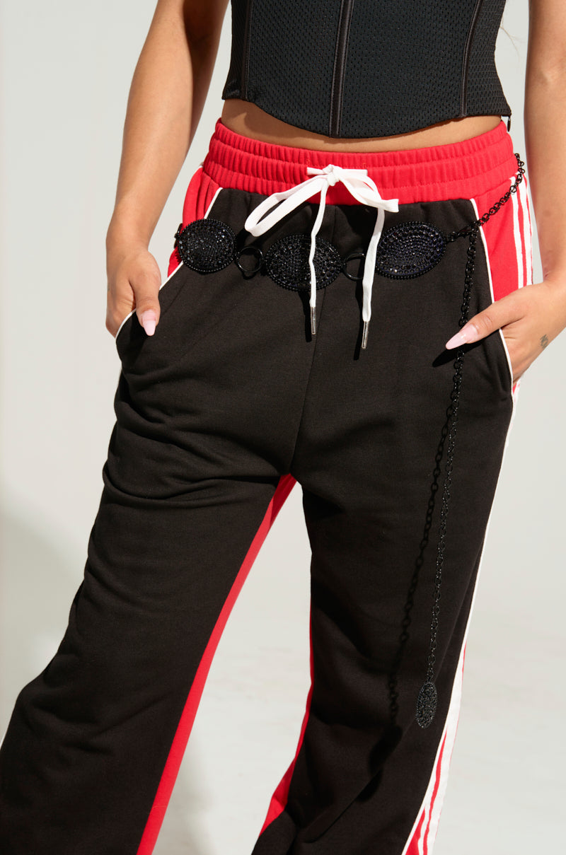 A person is wearing a black corset top paired with track pants that combine red and black hues, featuring white drawstrings. The track pants, highlighted by their decorative rhinestone elements, offer an adjustable fit and are accented with red and white stripes along the sides. Their hands rest casually in the pockets, subtly complementing the look with the CARTER BLING CHAIN BELT. 2