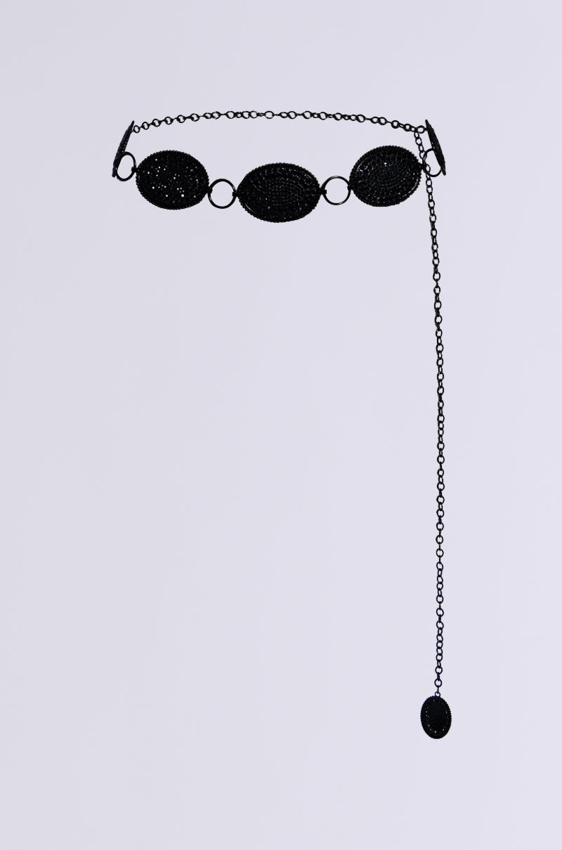 The CARTER BLING CHAIN BELT is a black accessory adorned with four large, interlinked oval links featuring rhinestone accents. It includes an extended chain that ends with a dangling oval piece, and its adjustable fit is highlighted against a gray background in the photograph. 1