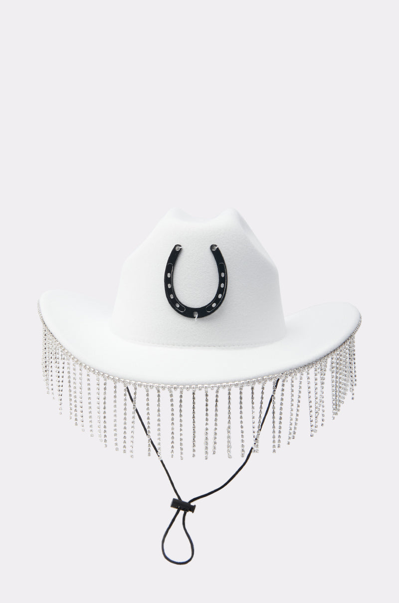 The TEST MY LUCK HORSESHOE HAT is a white Western hat with a black horseshoe on the front, shimmering rhinestone fringe along the brim, and a black adjustable chin strap.