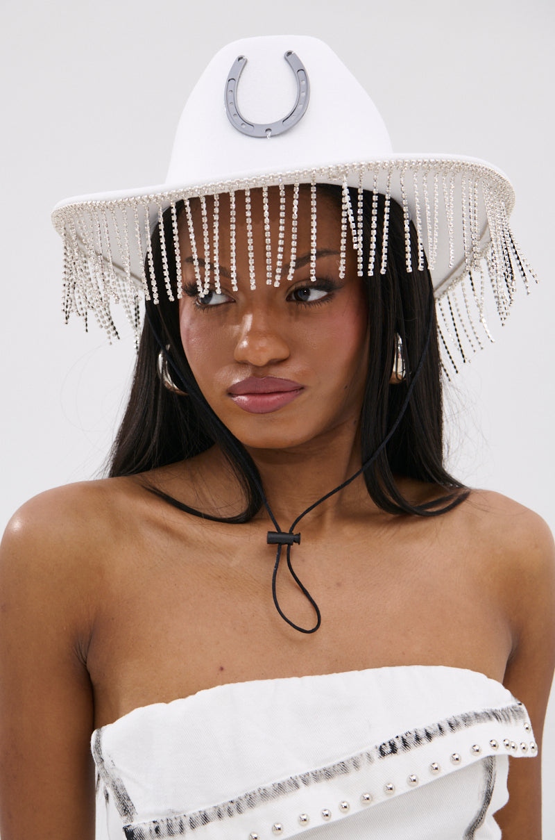 A woman wears the TEST MY LUCK HORSESHOE HAT, a white Western hat adorned with a horseshoe emblem and rhinestone fringe, secured under her chin. She pairs it with a white textured strapless top with metallic accents.