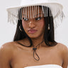 A woman wears the TEST MY LUCK HORSESHOE HAT, a white Western hat adorned with a horseshoe emblem and rhinestone fringe, secured under her chin. She pairs it with a white textured strapless top with metallic accents.