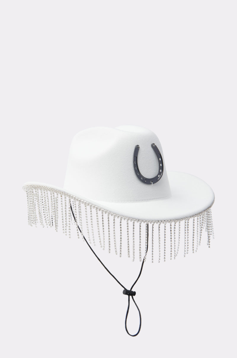 The TEST MY LUCK HORSESHOE HAT is a white Western hat with a black horseshoe emblem, black chin strap, and sparkling rhinestone fringe along the brim.