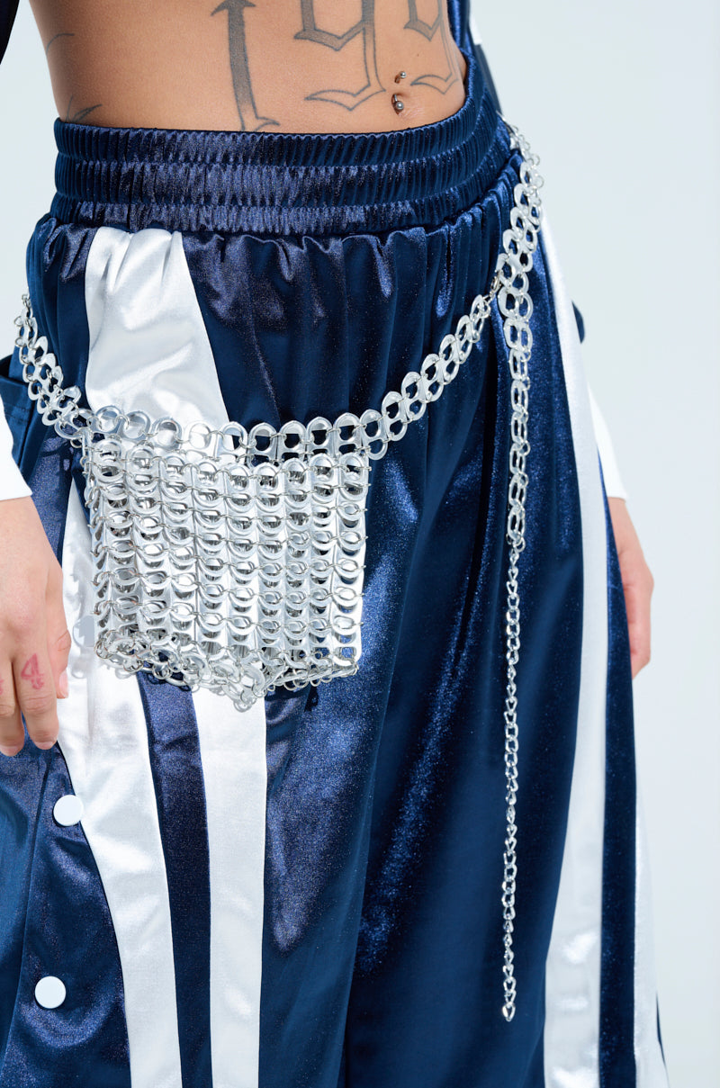 A person is wearing navy and white satin pants with an adjustable chain belt featuring the shiny silver WAYNE POP TAB BELT BAG, creating a unique look. A tattoo is partially visible on their torso. 2