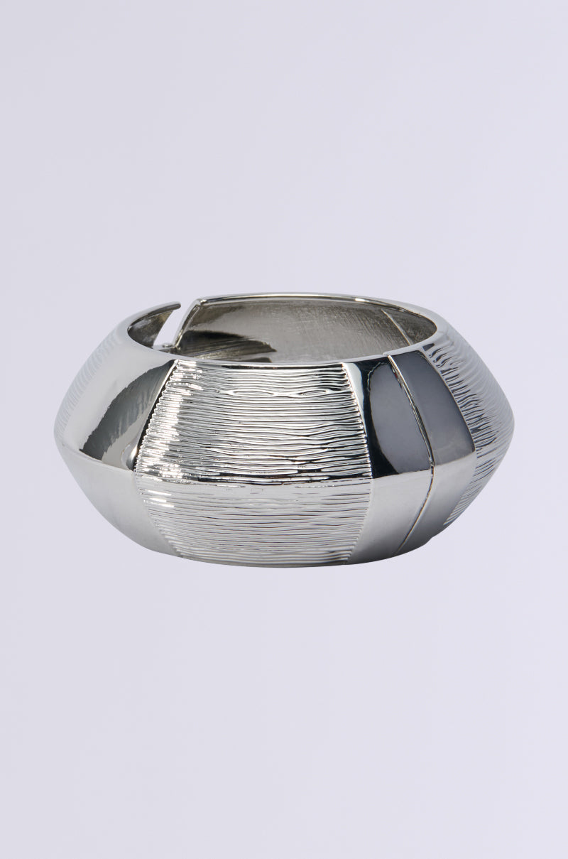 The OUT OF THIS WORLD CUFF is a statement bracelet featuring a silver bangle with a geometric design of alternating smooth and textured surfaces. Its chunky silhouette includes an open hinge, exuding an otherworldly charm.