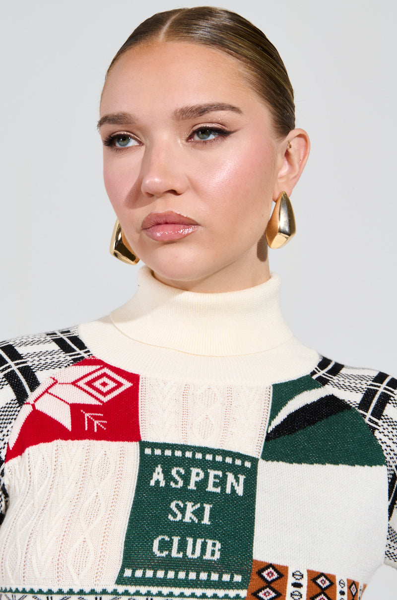 A person with slicked-back hair is wearing a colorful sweater adorned with the text "Aspen Ski Club" and various patterns, complemented by large metallic faux gold earrings, named the Zelly Statement Earrings. The background is plain and light-colored. 1