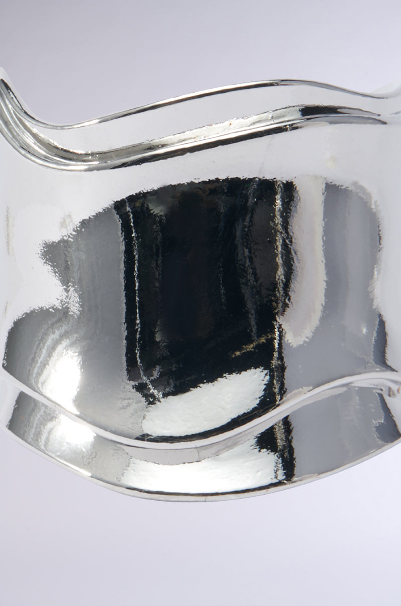 Close-up of a chic piece, the SISI Statement Bracelet Cuff in Silver, featuring abstract curved and reflective surfaces with distorted reflections. Set against a plain, soft gray background, this artistic marvel emphasizes its faux silver metallic texture and flowing lines. 3