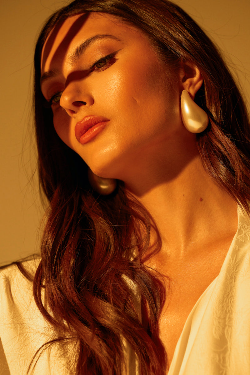 A woman with long brown hair and a thoughtful expression looks upward, her serene visage accentuated by sunlight casting gentle shadows. Her PEARLESCENT VIBES STATEMENT EARRING adds elegance to her look, harmonizing with the warm, golden hue of the scene as she wears a white top.