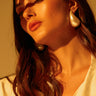 A woman with long brown hair and a thoughtful expression looks upward, her serene visage accentuated by sunlight casting gentle shadows. Her PEARLESCENT VIBES STATEMENT EARRING adds elegance to her look, harmonizing with the warm, golden hue of the scene as she wears a white top.