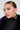 A person with slicked-back hair and large, square-shaped THE BIGGER THE HOOP EARRING is wearing a black ribbed turtleneck and a dark fur-like coat. The person gazes to the side with a neutral expression against a plain background.