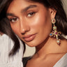 A woman with long dark hair and smooth skin wears a white top and the YOUR MAJESTY EARRING, a large cross-shaped earring embellished with rhinestones and colorful gemstones, gazing into the camera with confidence.
