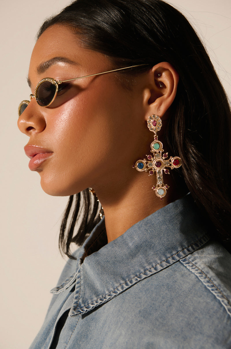 A person wearing oval sunglasses and YOUR MAJESTY EARRING, adorned with colorful rhinestones in a cross shape. They pair the earrings with a denim collared shirt as lighting accentuates their facial features against a neutral backdrop.