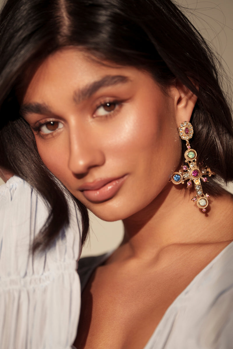 A woman with long dark hair and smooth skin wears a white top and the YOUR MAJESTY EARRING, a large cross-shaped earring embellished with rhinestones and colorful gemstones, gazing into the camera with confidence. 0