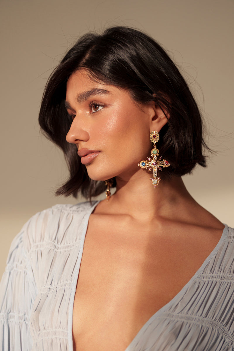 A woman with short dark hair wears the YOUR MAJESTY EARRING—large, rhinestone-encrusted cross earrings with colorful gems—and a light, textured deep V-neck blouse, gazing to the side against a neutral background. 1