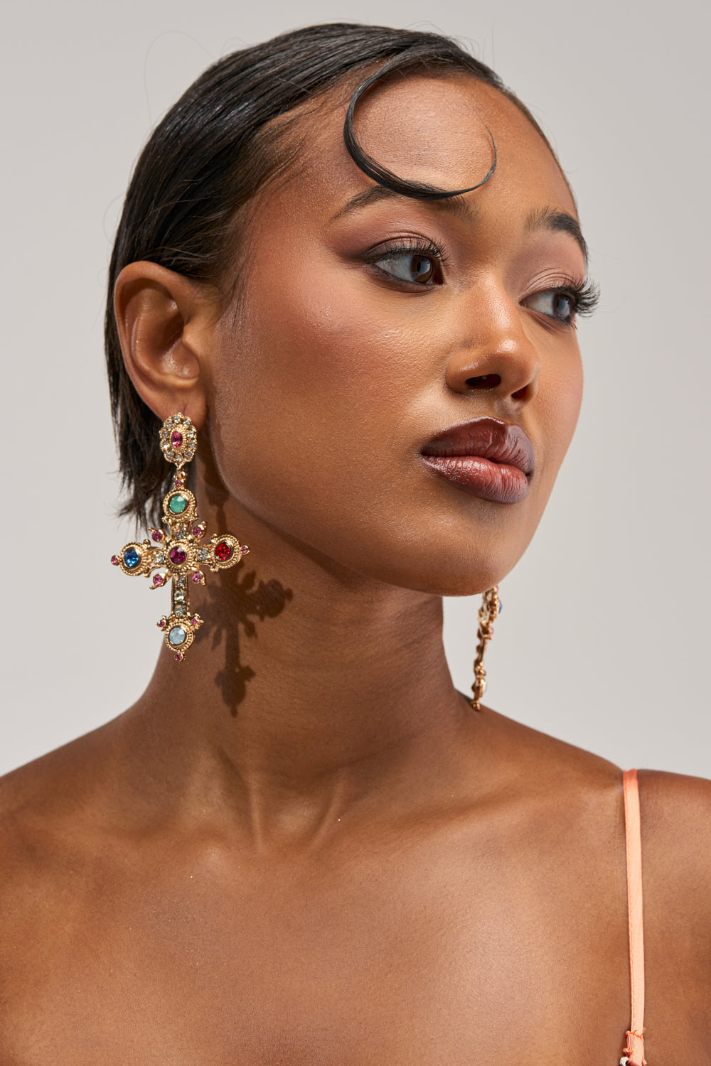 A woman with short, styled hair wears the YOUR MAJESTY EARRING—a large, ornate gold cross adorned with colorful gems. She has glowing, natural makeup and gazes to the side against a neutral background.