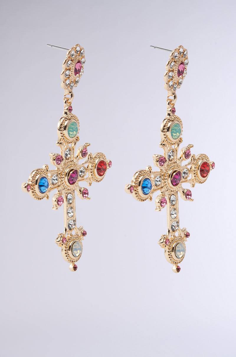 The YOUR MAJESTY EARRING by AKIRA features gold cross-shaped earrings with rhinestone-adorned multicolored gemstones in blue, green, red, and clear hues. Each intricate piece comes with a hook for wearing against a neutral gray backdrop.