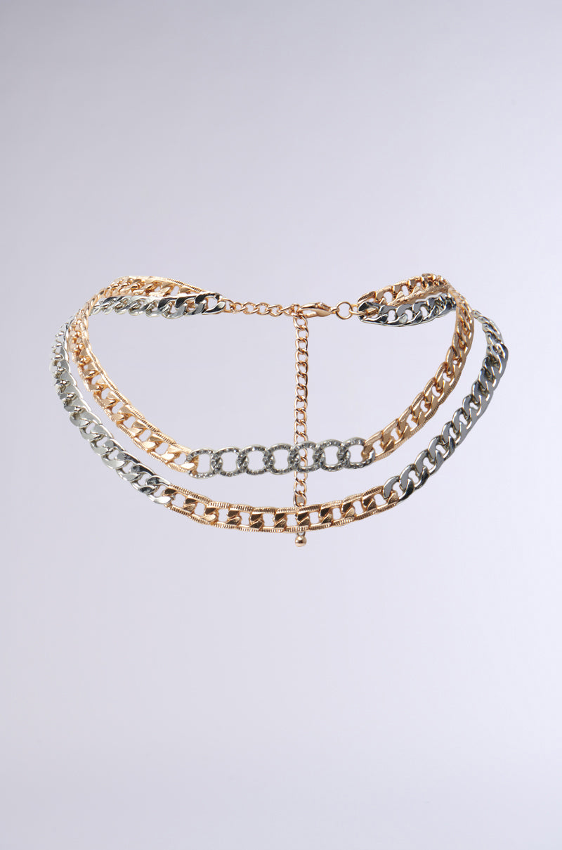 Introducing the LAYERED UP NECKLACE: a chic accessory with a blend of gold and silver Cuban chain links, featuring a layered design and a rhinestone-embellished centerpiece. This stylish and modern necklace stands out beautifully against a plain gray background.